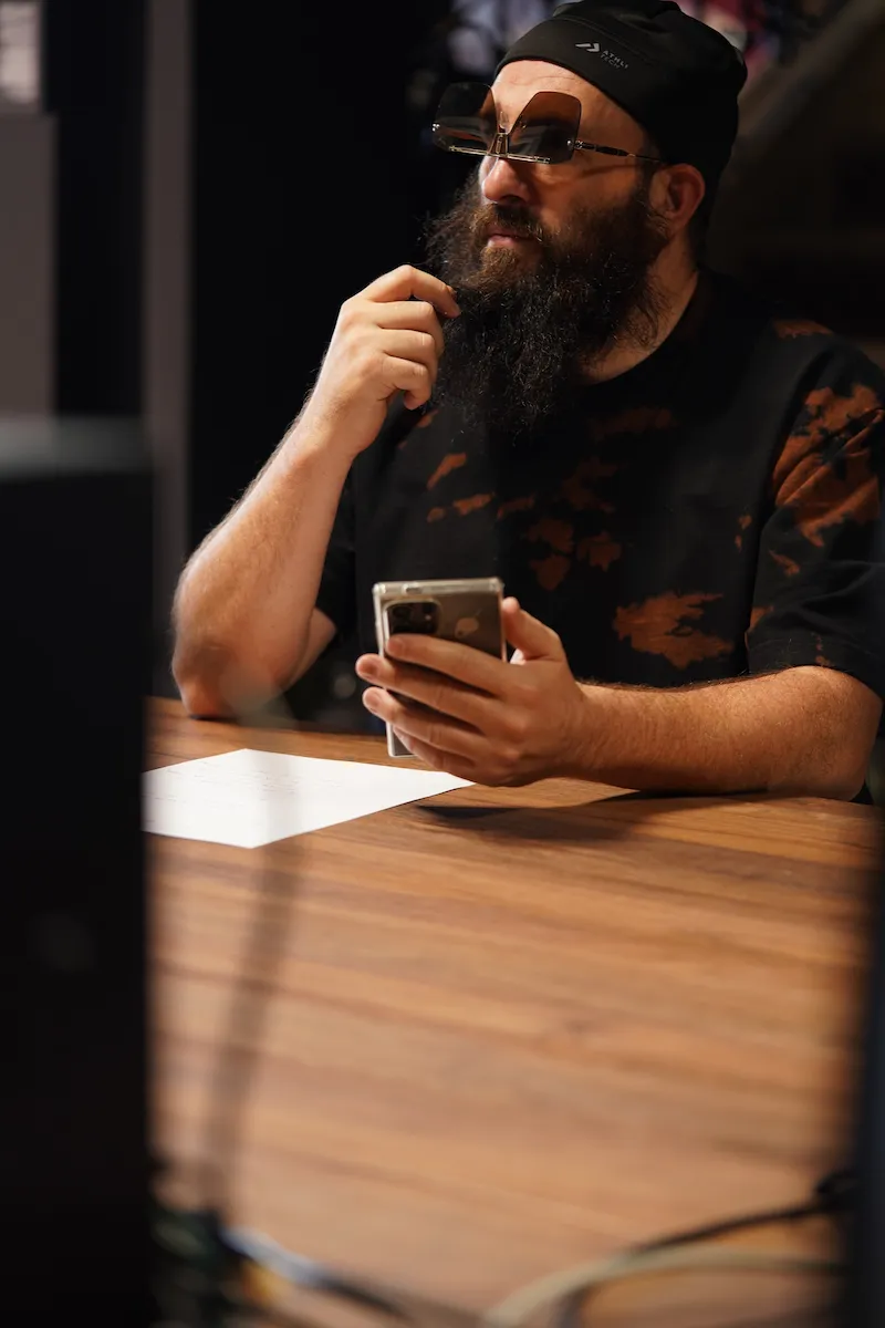 Homme barbu avec lunettes design et smartphone en main, style créatif et concentré Homme barbu portant des lunettes carrées originales et un bonnet noir, assis à une table avec un smartphone