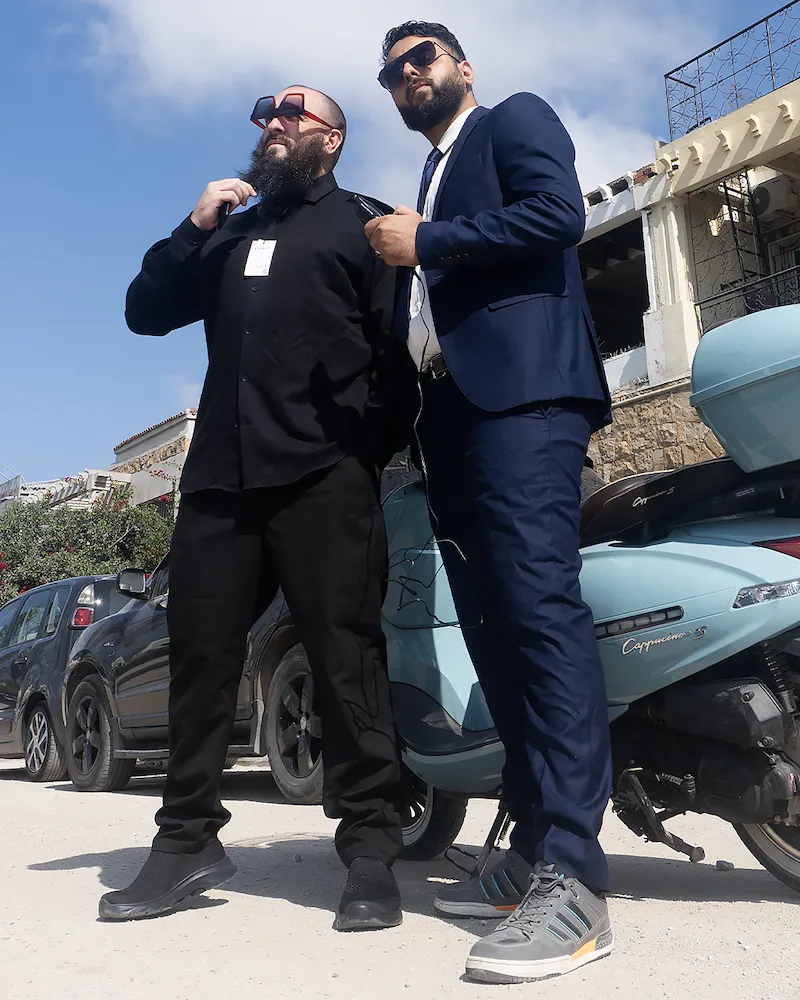 Superfoodman et son faux garde du corps en tenue d’agents secrets devant un scooter et des voitures